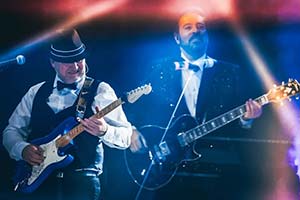 Zwei Gitarristen nebeneinander im blauem Bühnenlicht. Linker Gitarrist mit Hut.